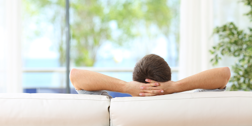 Man relaxes on white couch in living room and considers benefits of ductless vs central air conditioning.