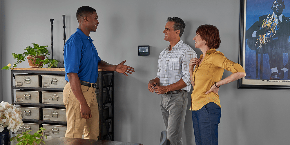 Tech in blue shirt shows homeowning couple HVAC troubleshooting tips in their home.