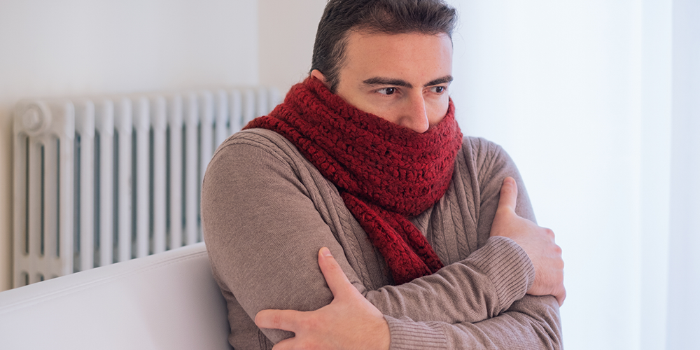 Too cold at home, a man sits bundled up on couch in front of radiator.