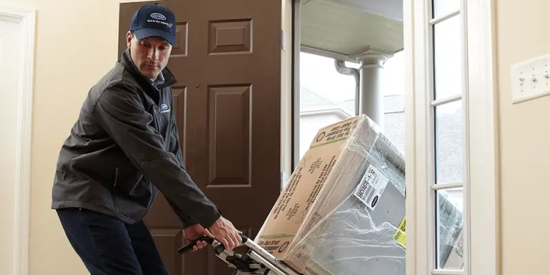 HVAC tech in dark jacket and hat wheels a new furnace installation into residential home.