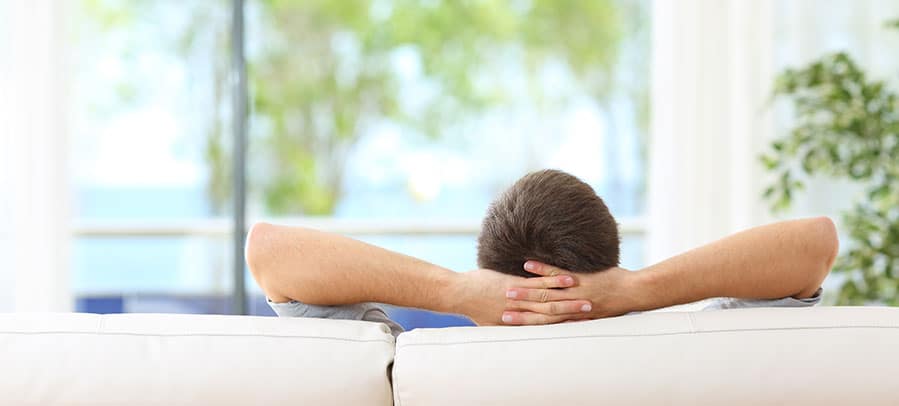 Air Quality Male homeowner relaxes on white living room couch with hands behind his head.