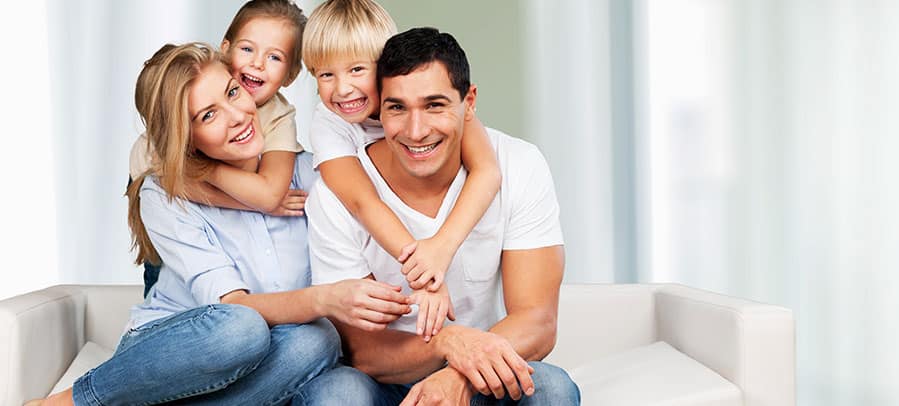 Mother, father, and two young children all dressed in white shirts smile for the camera on their living room couch.