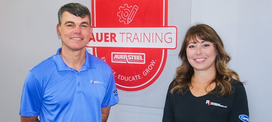 The Story Behind RJ Heating RJ owners Julius and Erin smile in front of Auer Steel Training plaque inside training lab.